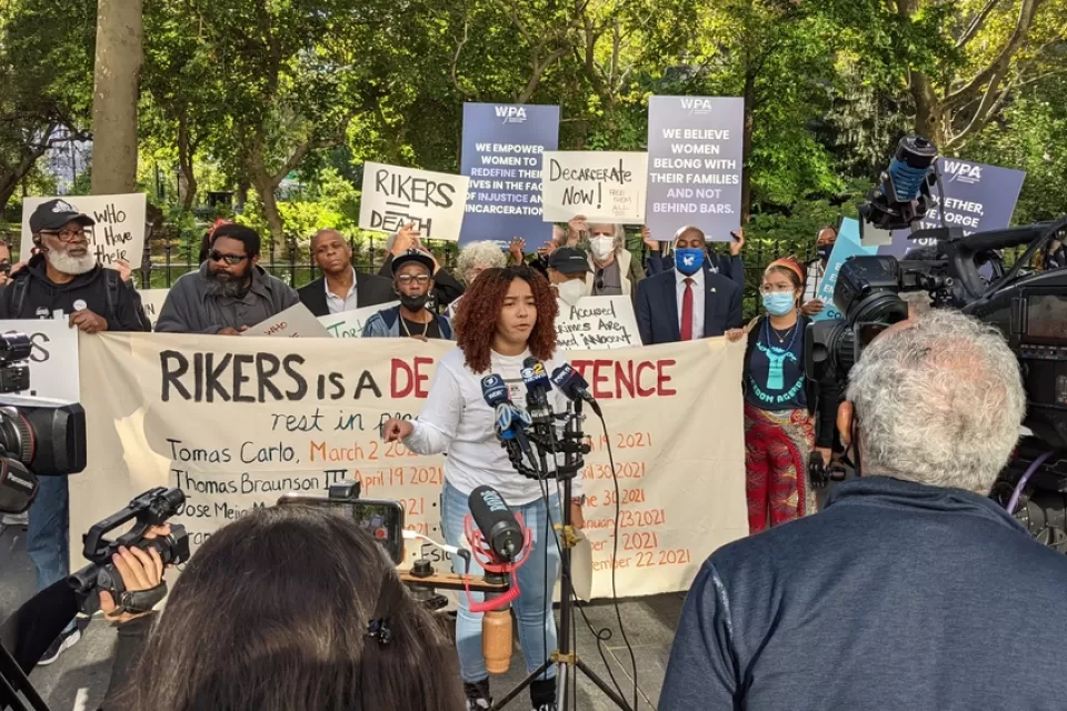 Crowd of activists protesting conditions at Rikers Island with one woman in front speaking into microphone